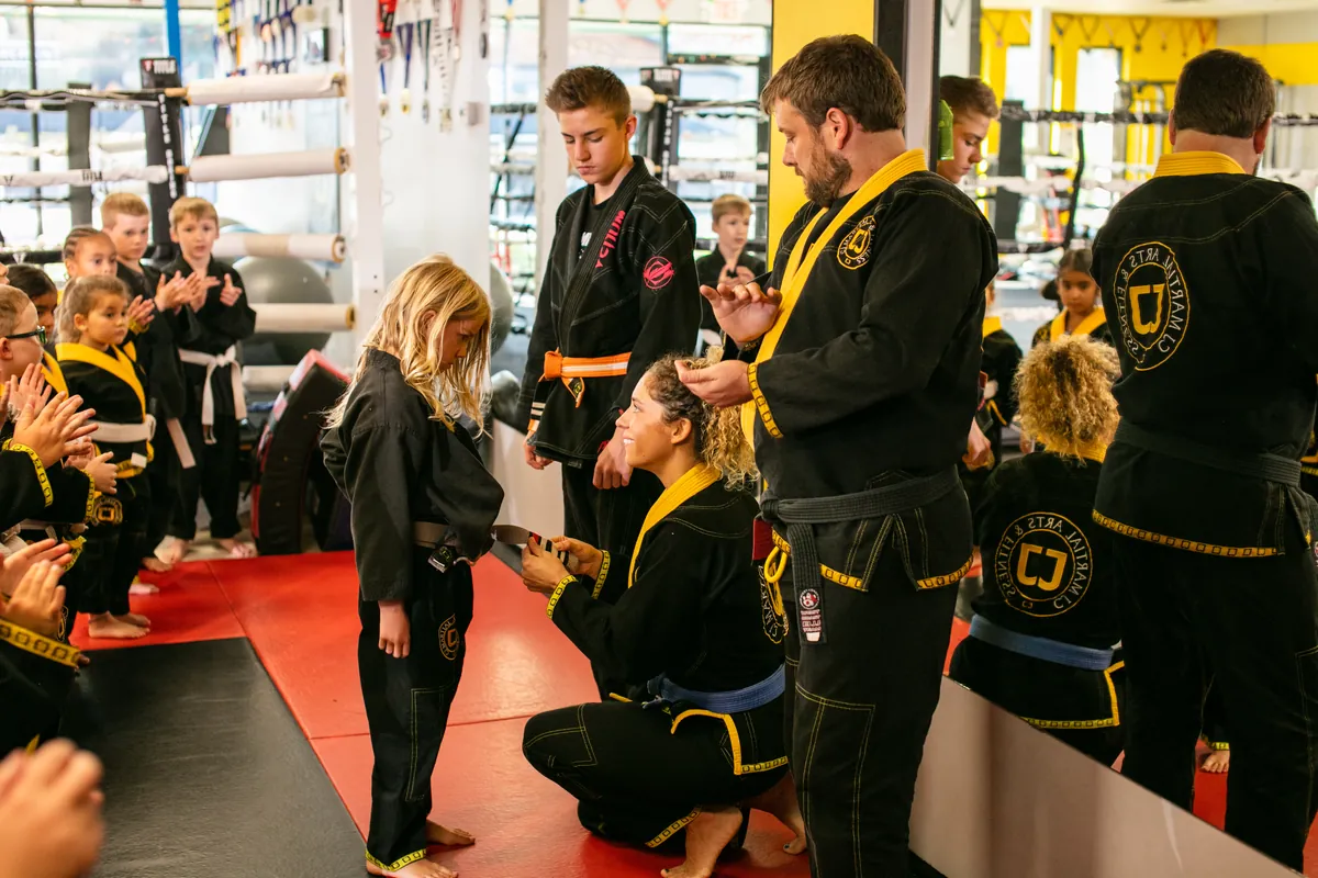 A young student receiving a new belt from CJ Martial Arts instructors while classmates applaud